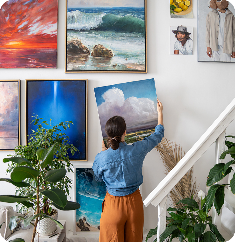 A woman hangs a landscape painting in a cozy, art-filled living space.